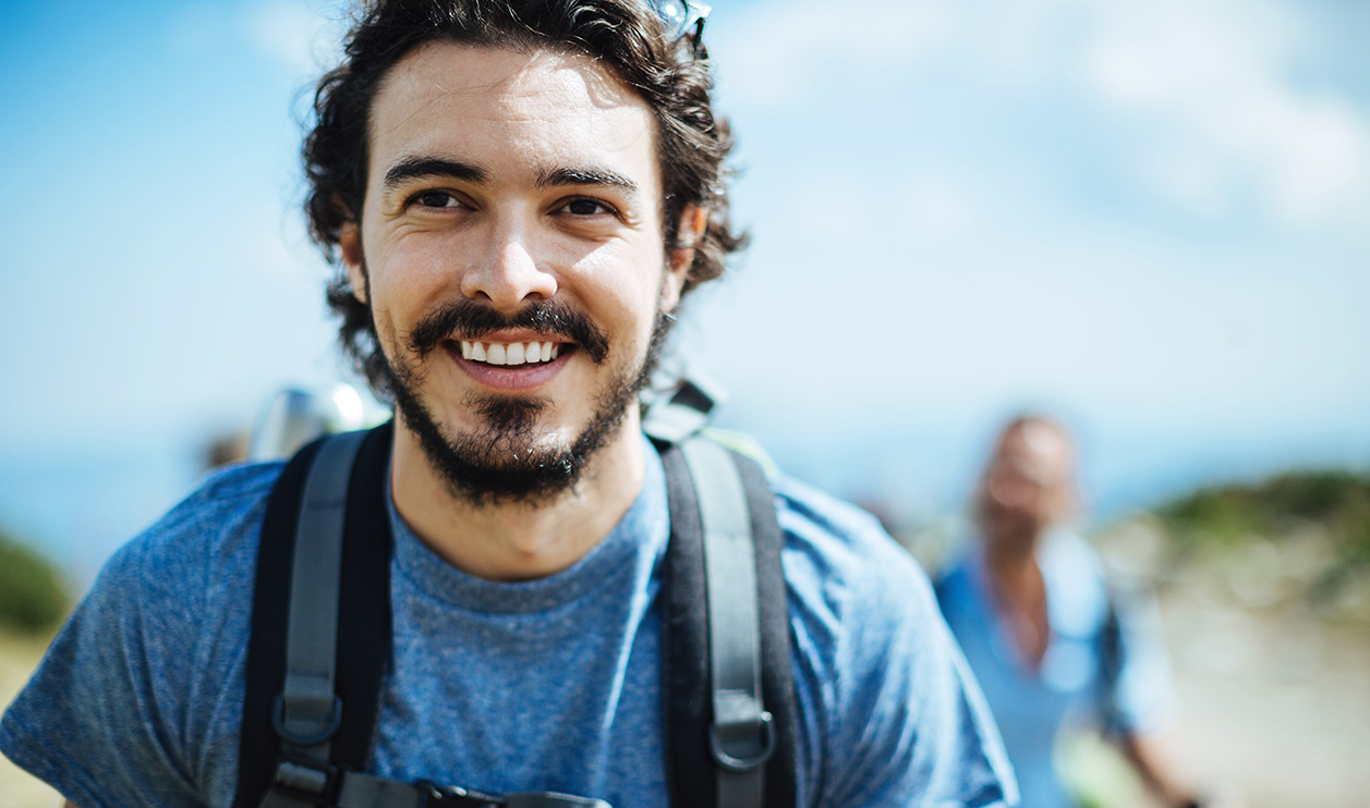 Wandernder Mann mit Rucksack lächelt vor blauem Himmel.